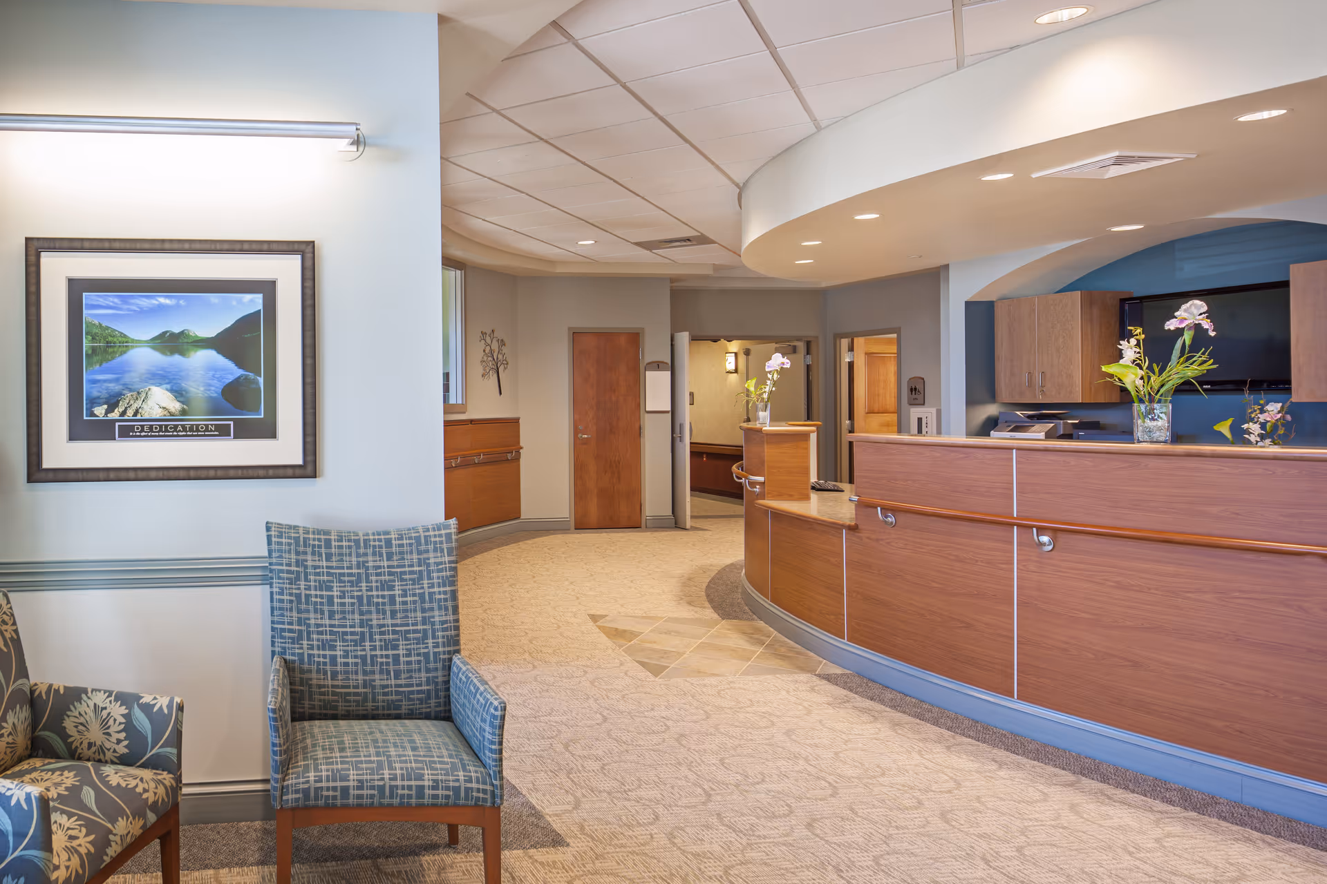 Reception area of Andover Forest Post Acute Care Center featuring a curved wooden front desk with flowers in vases, a blue patterned chair, a floral armchair, a framed landscape photograph on the wall, and a hallway with wooden doors in the background.
