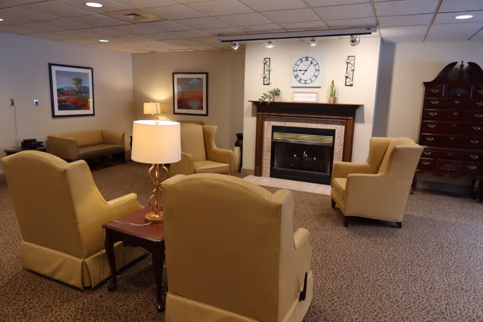Cozy lounge with beige armchairs, a fireplace, and framed art in a senior living facility.