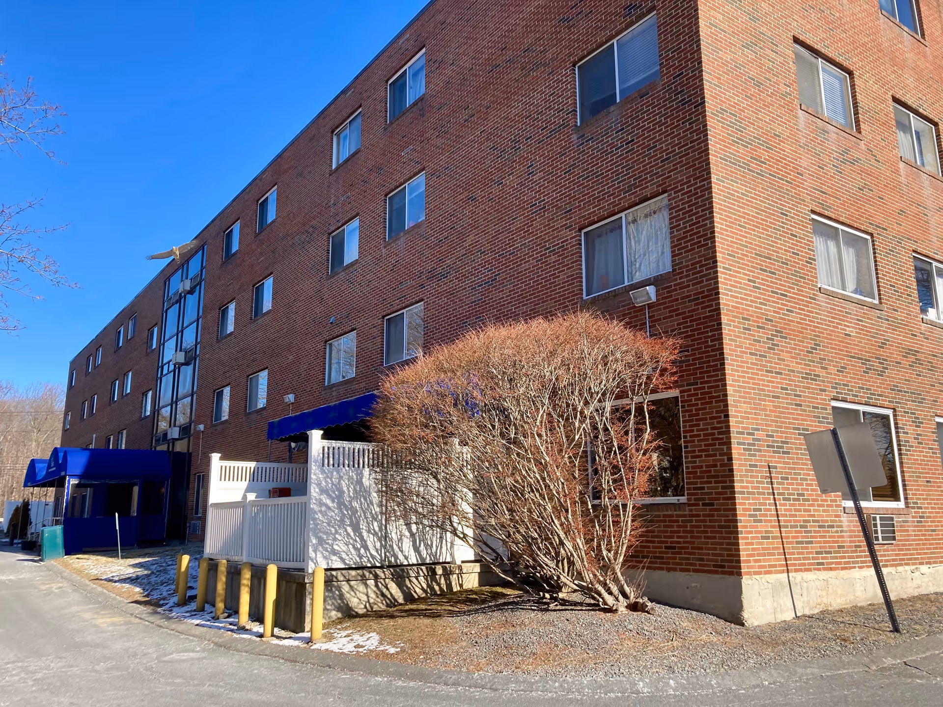 Brick multi-story building with blue awnings at the entrance, bare shrubs, and a clear blue sky.