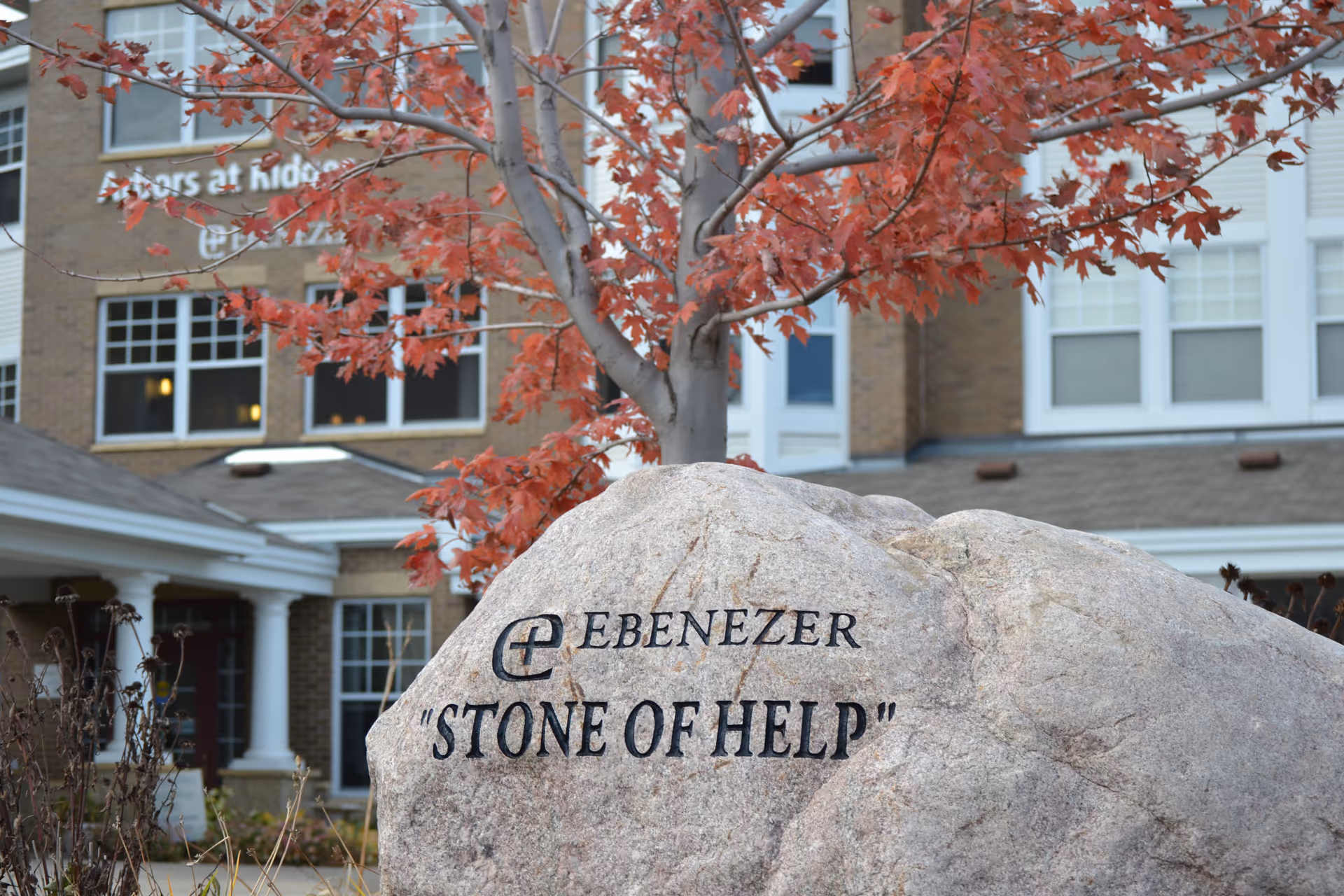 A rock engraved 'Stone of Help' sits before a brick senior living building with red leaves.