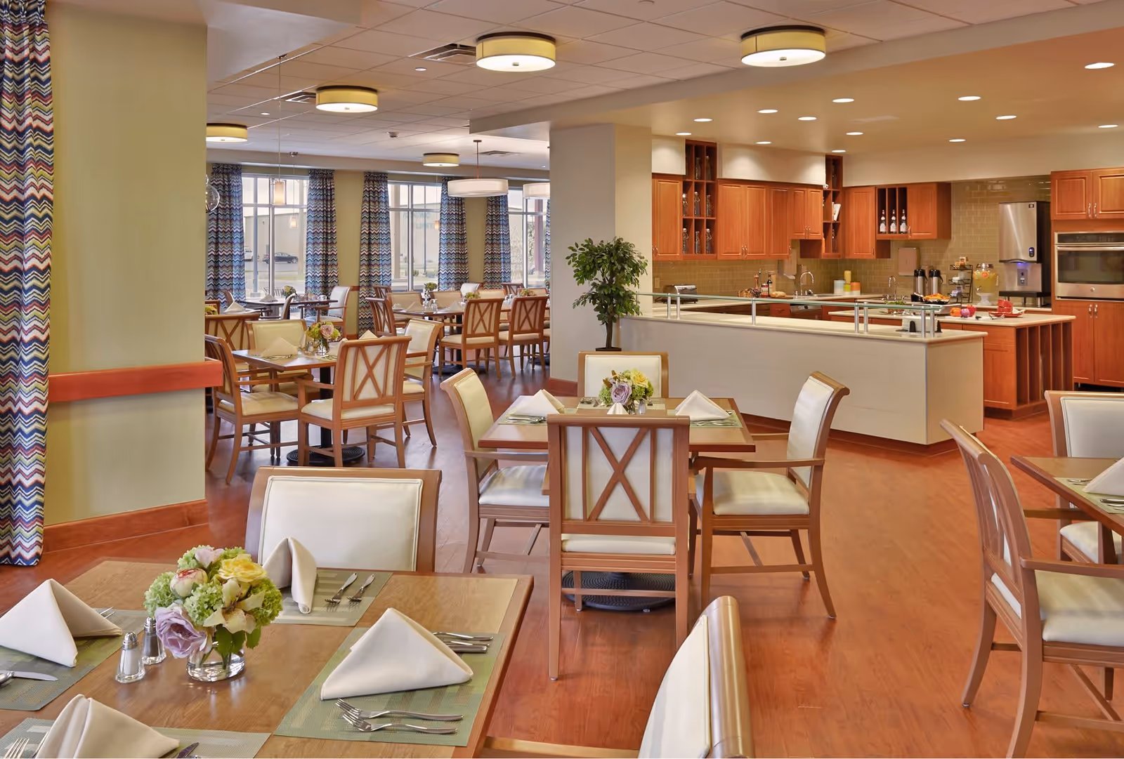 Bright dining room with neatly set tables, floral centerpieces, and an open kitchen in the background.