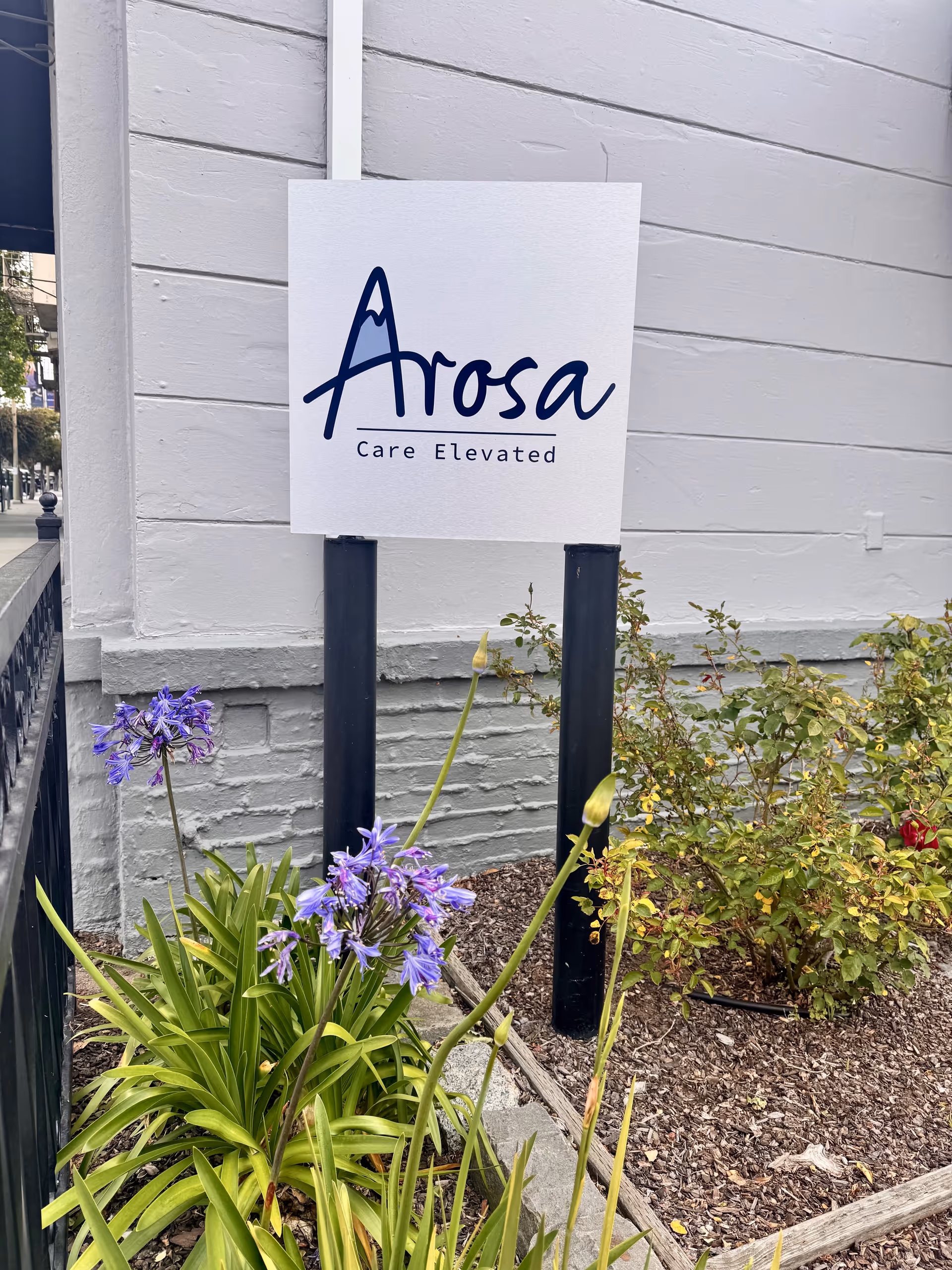 Arosa care sign mounted outside a light-colored building with purple flowers nearby