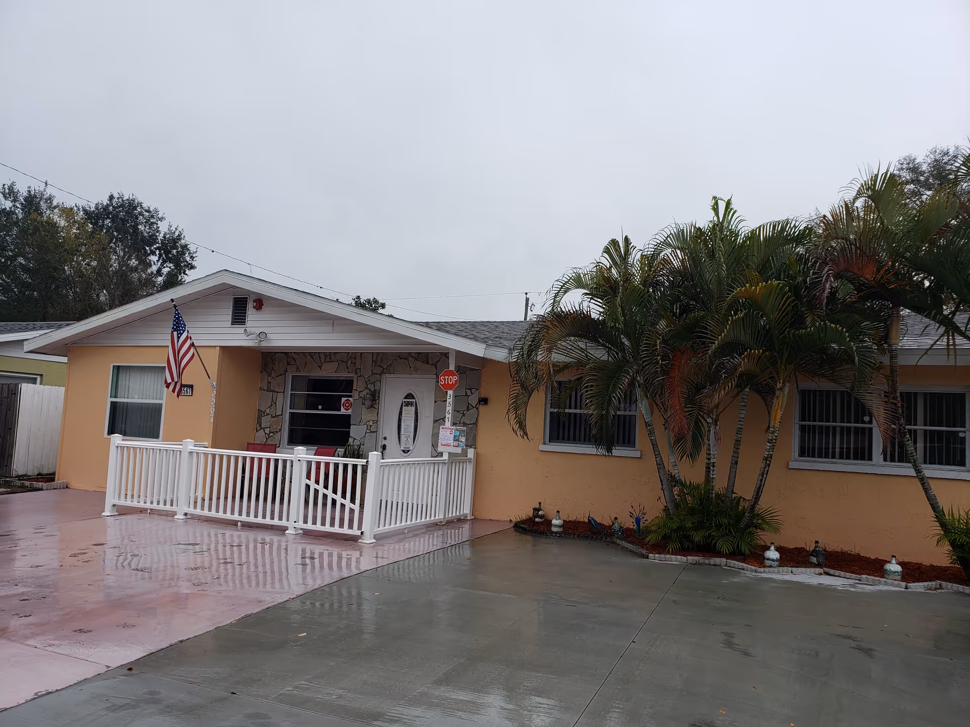 Peach-colored, single-story assisted living facility with a white railing, flag, and palm trees.