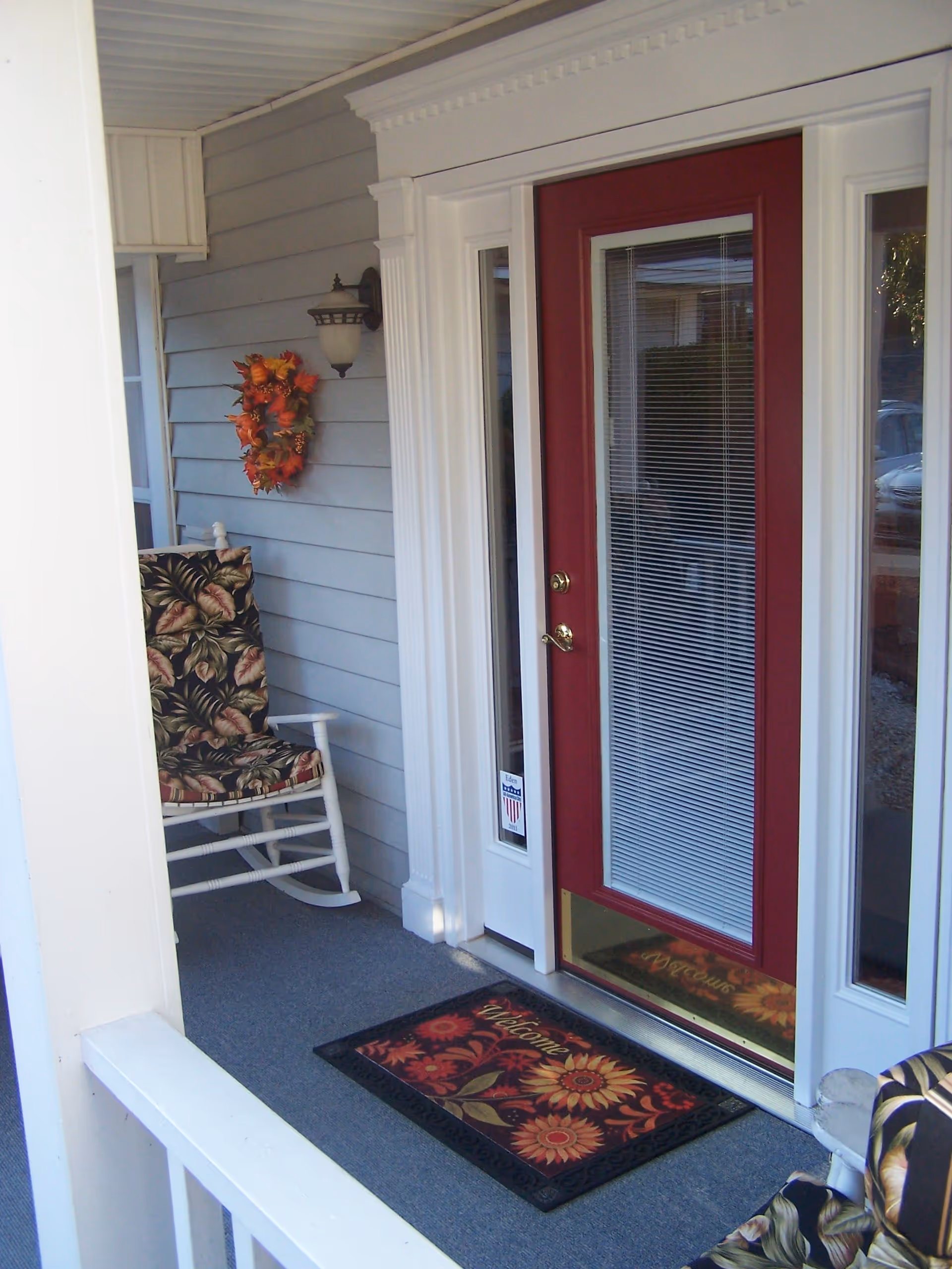A covered porch area with a red door featuring glass panels and white trim. There is a welcome mat with a floral design in front of the door. A white rocking chair with a floral cushion is placed on the porch, and a fall-themed wreath hangs on the gray siding wall next to a wall-mounted light fixture.