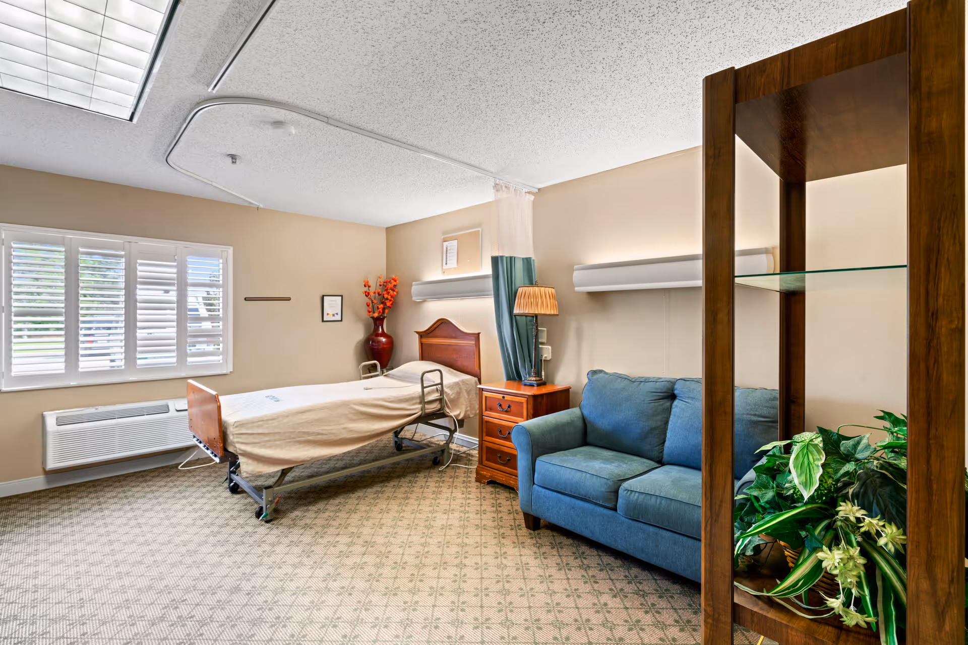 A well-lit room in a healthcare facility featuring a hospital bed with beige bedding, a wooden headboard, and a side table with a lamp. There is a blue upholstered loveseat next to the bed, a large window with white shutters, a red vase with orange flowers, and a wooden shelving unit with green plants. The room has beige walls and patterned carpet flooring.