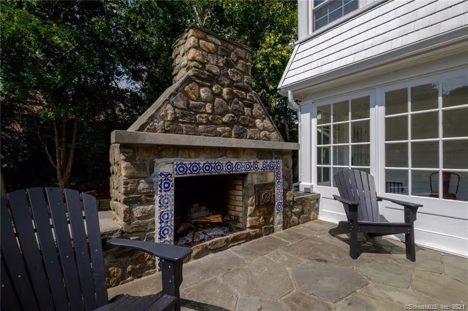 Stone outdoor fireplace with blue-tiled trim beside a white house and patio chairs.