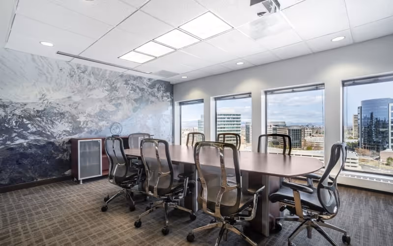A modern conference room with a long table, multiple chairs, and city-view windows.