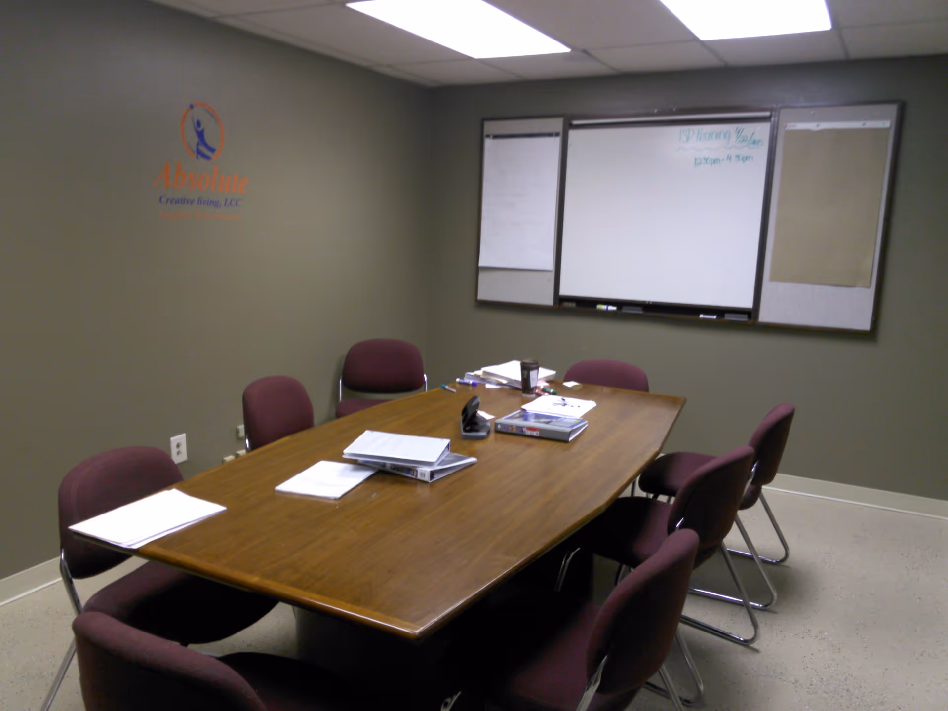 Meeting room with a wall logo, long table, chairs, papers, and a whiteboard.