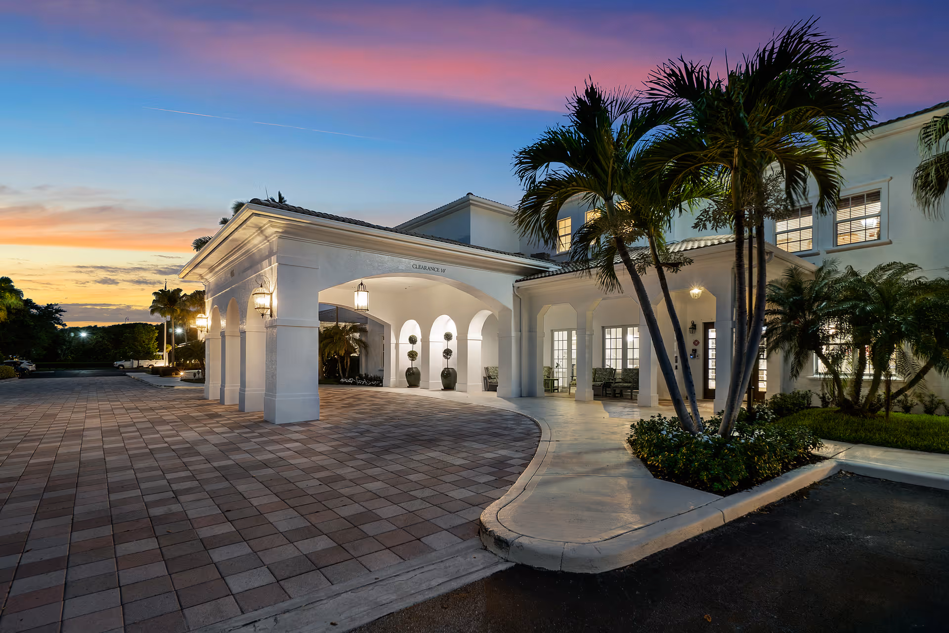 White Mediterranean-style building entrance with arches, palm trees, and a paved driveway at sunset.
