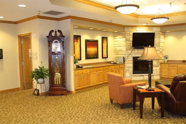 Warmly lit common room with upholstered chairs, a grandfather clock, built-in cabinets, a stone fireplace with wall-mounted TV, and framed artwork.