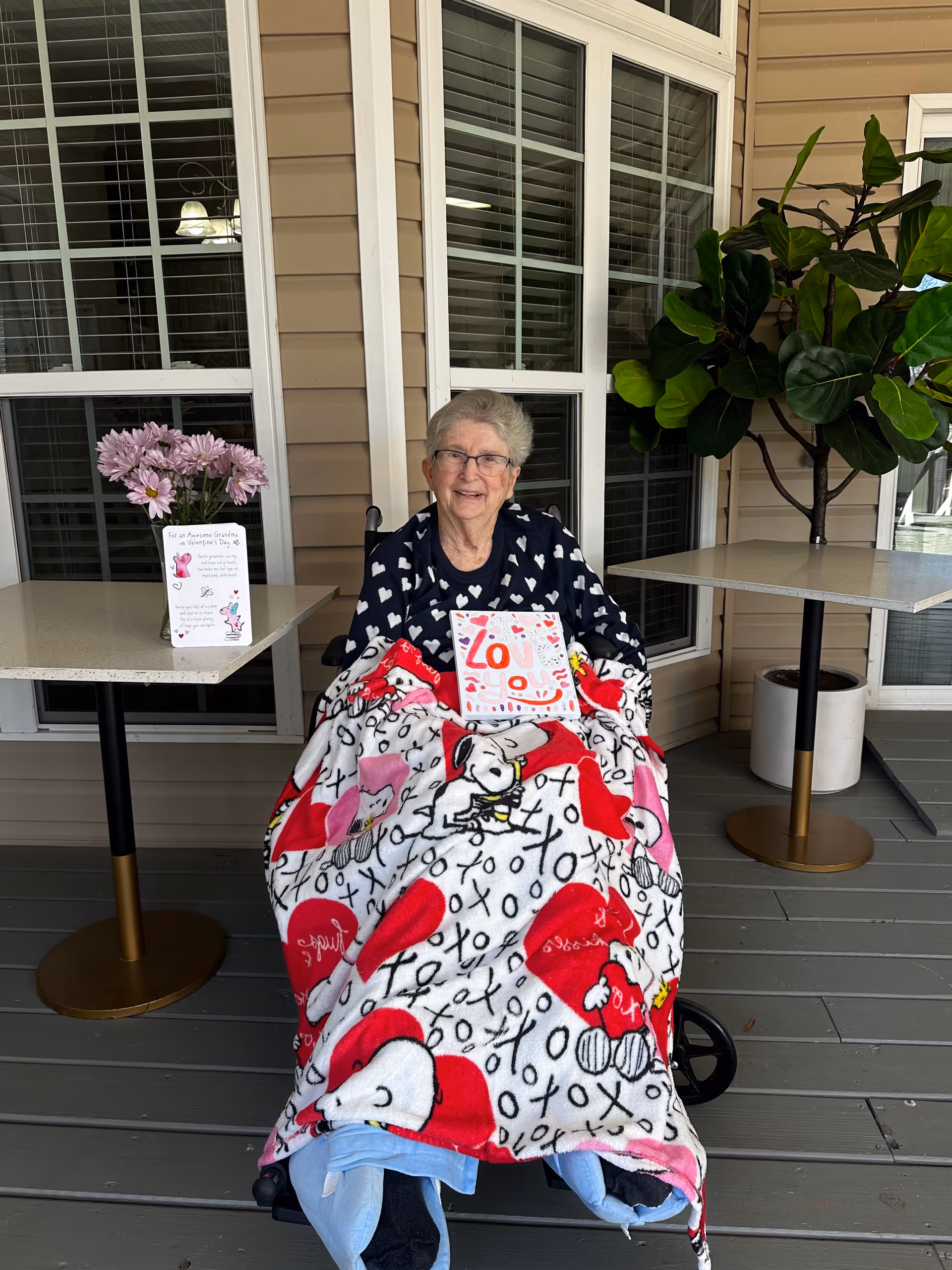 Elderly woman in a wheelchair on a covered outdoor patio with tables and plants.