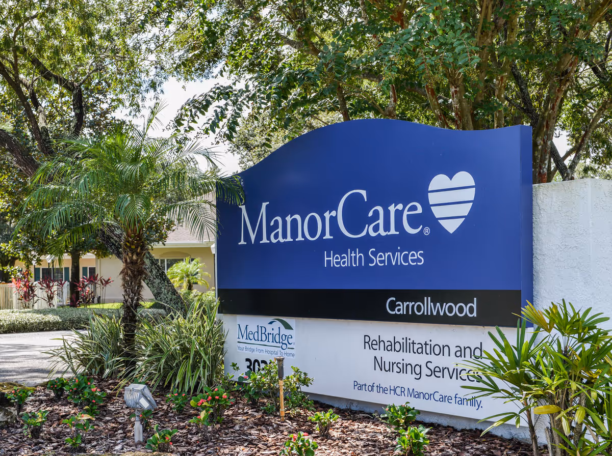 A large blue ManorCare sign with surrounding plants outside a rehabilitation facility.