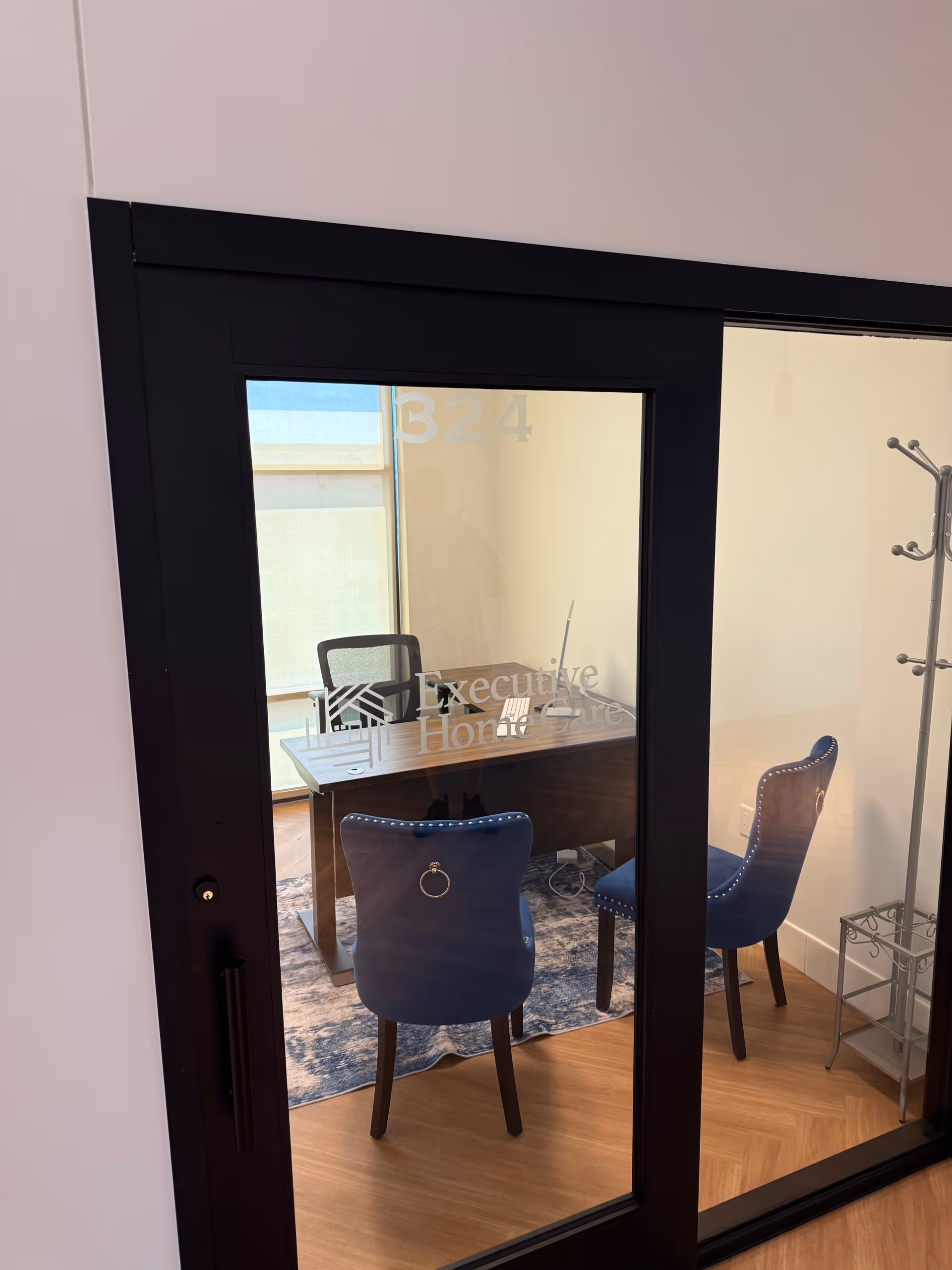 Glass door office with desk, two blue chairs, rug, and coat rack inside a modern room.