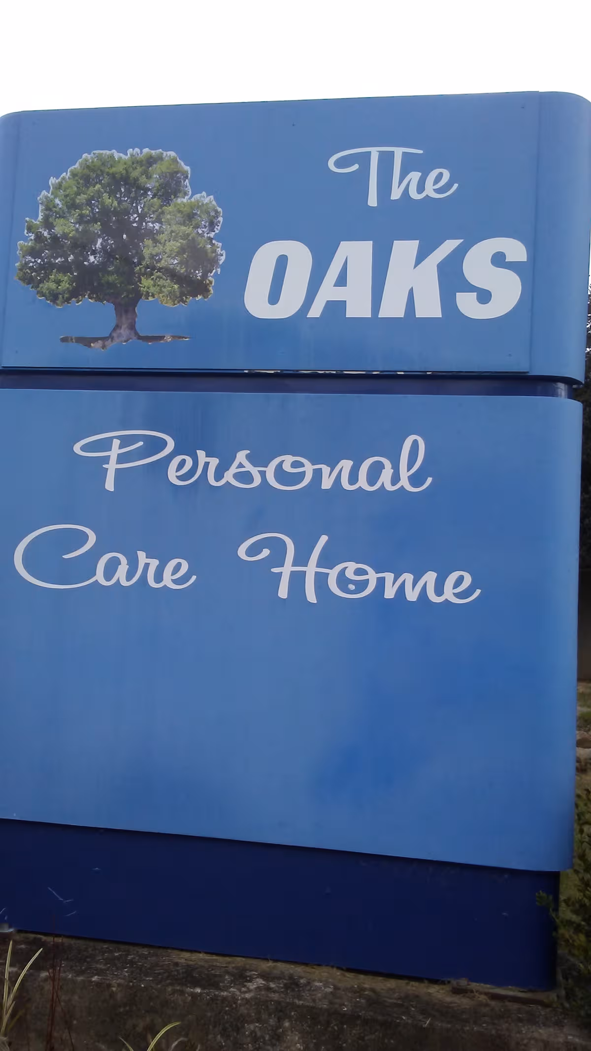 Blue outdoor sign reads The Oaks Personal Care Home with a tree graphic.