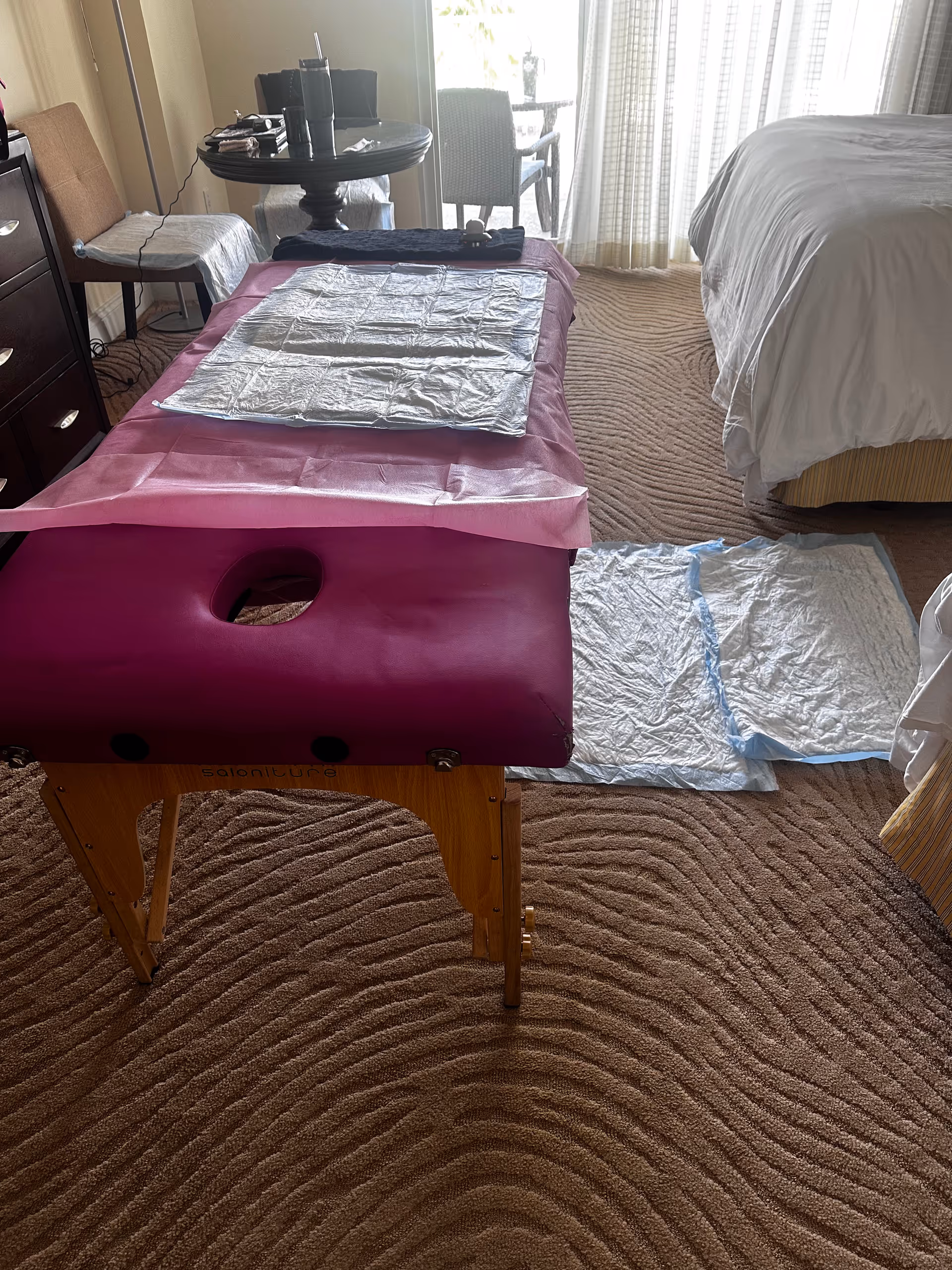 A pink massage table set up beside a bed in a hotel-like room.
