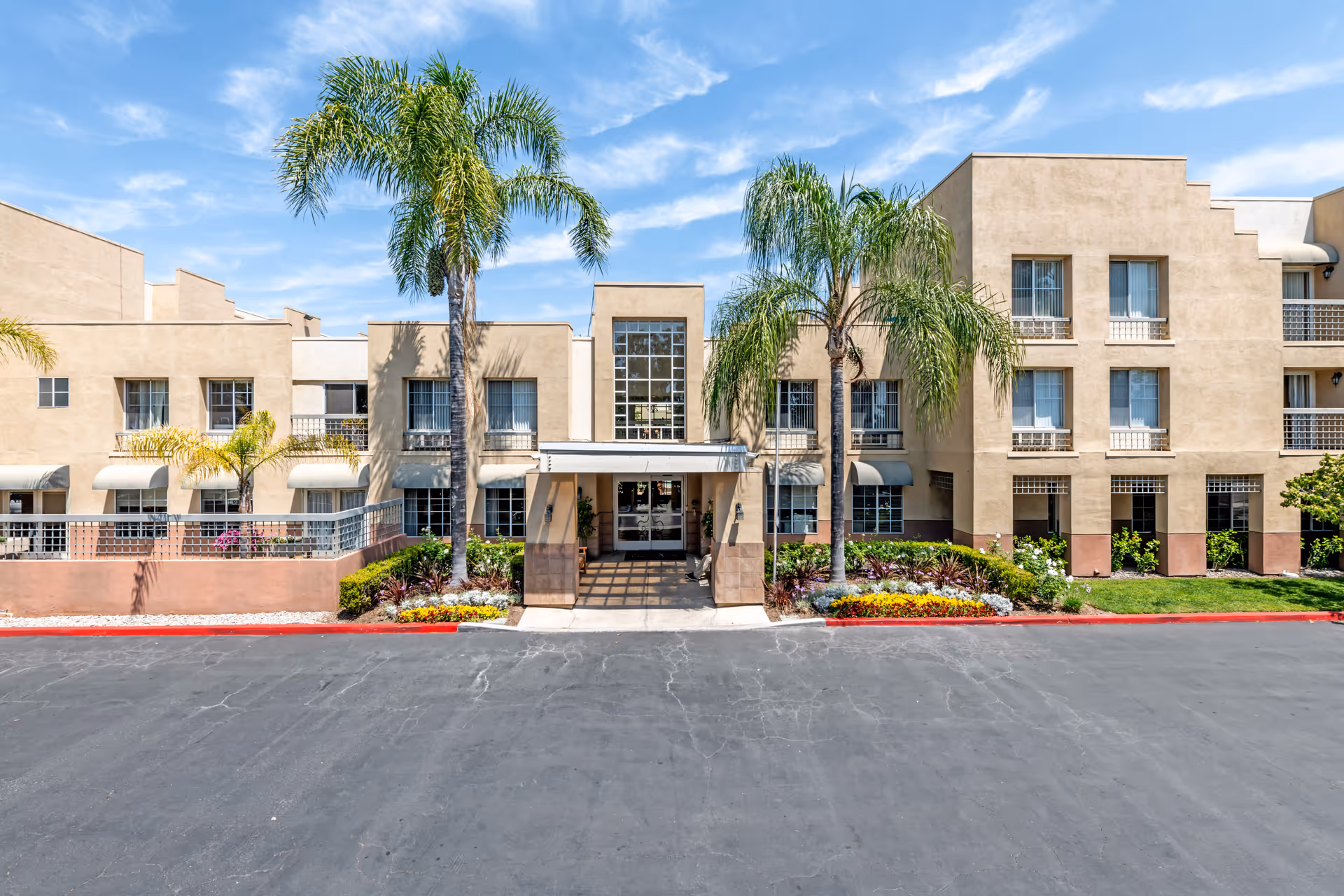 Exterior view of a beige multi-story senior living facility building named Park Plaza, with palm trees and landscaped flower beds in front, under a blue sky with scattered clouds.