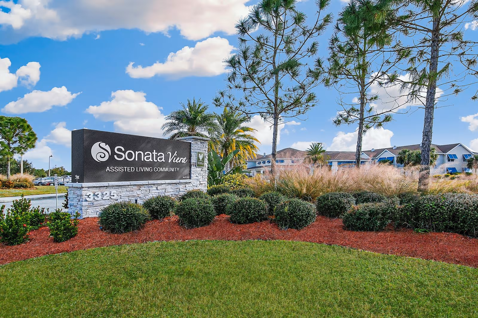 Outdoor view of the entrance sign for Sonata Viera Assisted Living Community surrounded by landscaped bushes, trees, and grass under a partly cloudy blue sky.