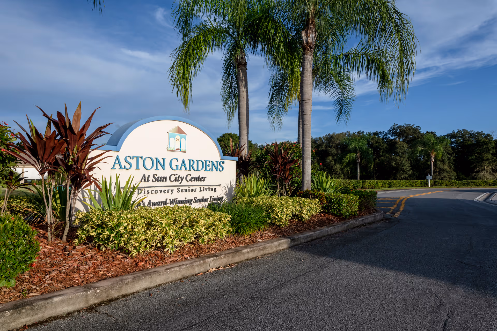 Entrance sign for Aston Gardens At Sun City Center surrounded by landscaped plants and palm trees under a blue sky.