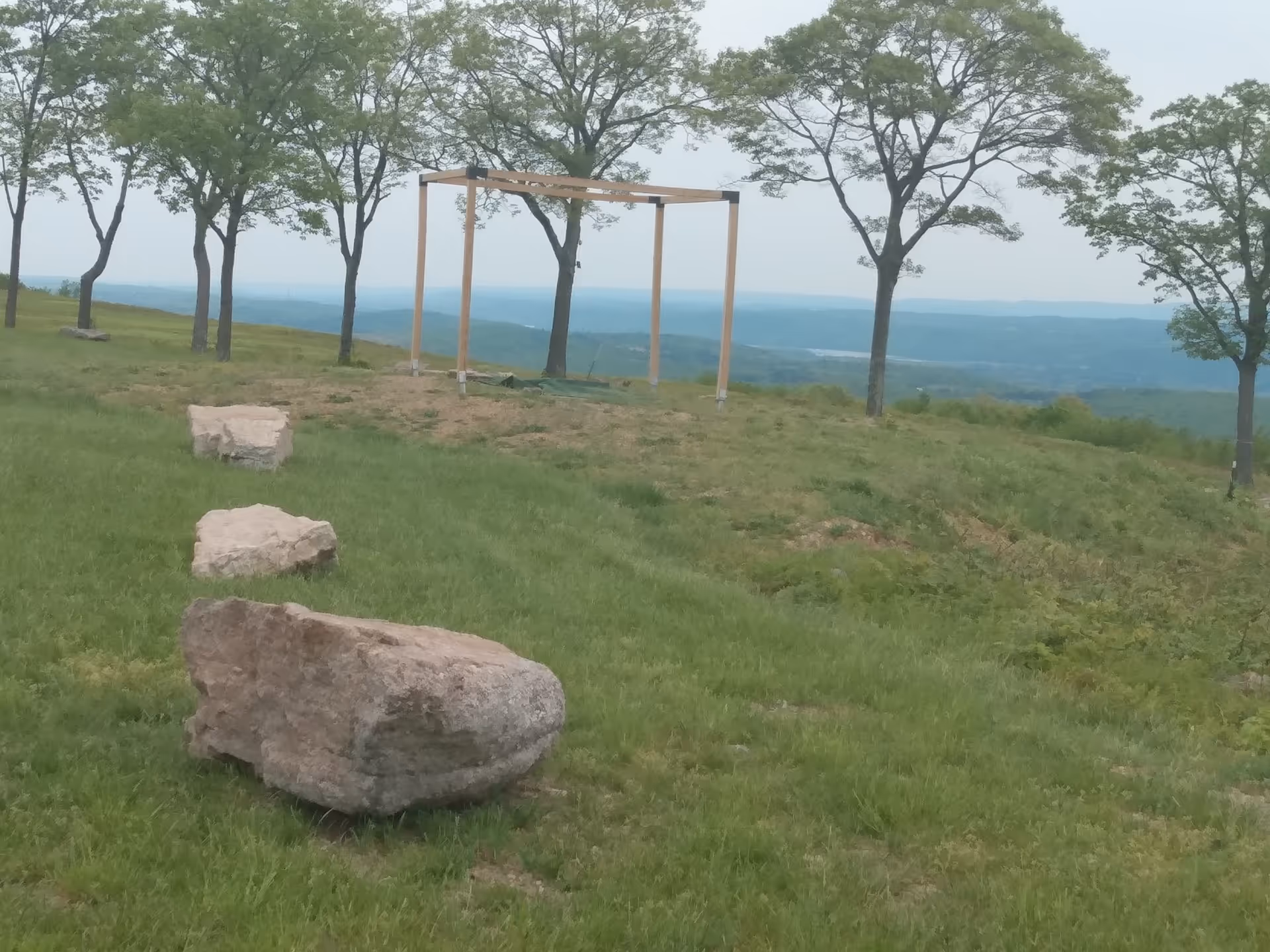 Open grassy hillside with rocks, trees, and a wooden pergola overlooking the horizon.