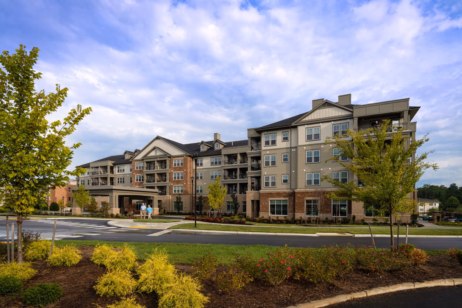 Exterior view of a multi-story retirement community building with a mix of brick and siding facade, surrounded by landscaped greenery and trees under a partly cloudy sky. Two people are walking near the entrance of the building.