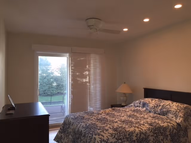 Bedroom at Tranquil Gardens senior living facility with bed, dresser, lamp, and balcony doors.