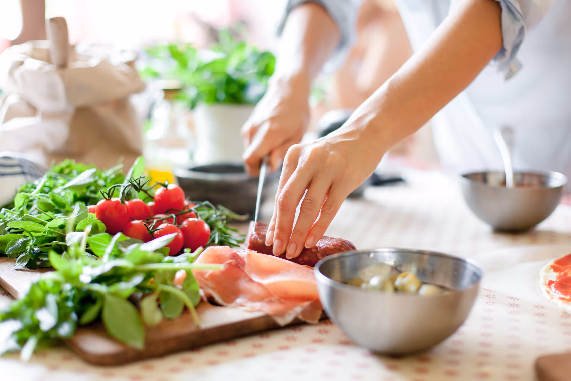 People prepare food in a bright kitchen, chopping herbs and slicing meat on a cutting board.