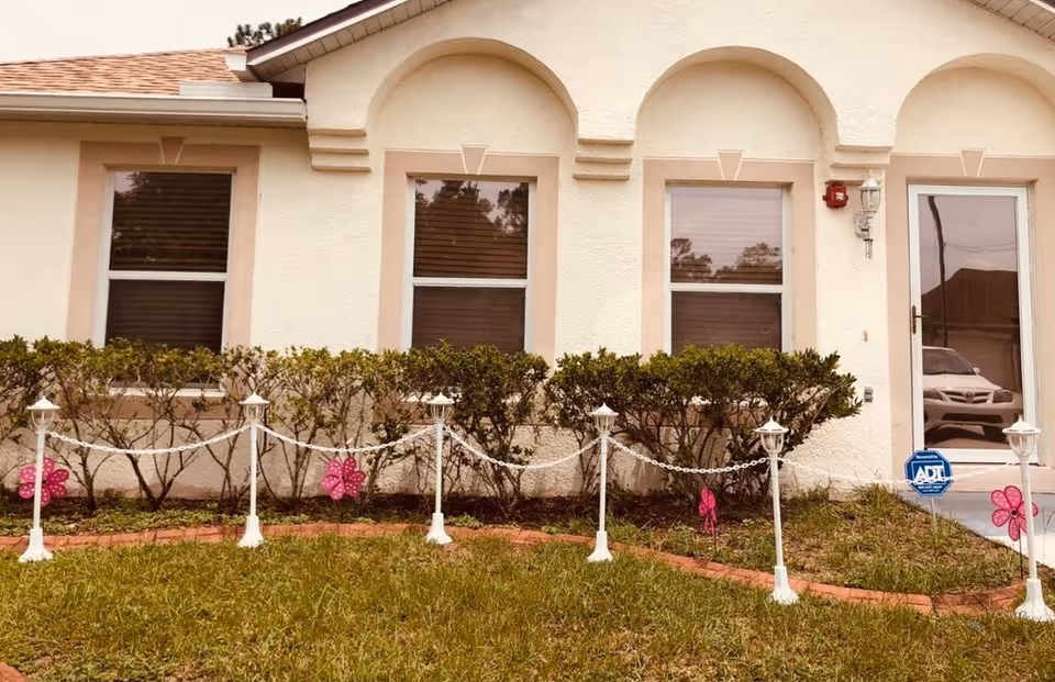 Front exterior of a beige senior living home with shrubbery, white posts, and a glass door.