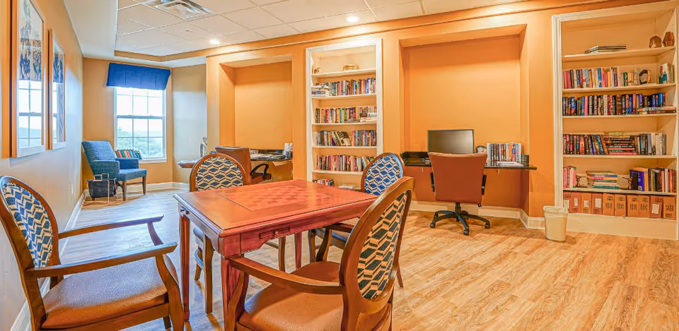 A well-lit room with a wooden table and four upholstered chairs in the center, a computer workstation with a chair against the wall, built-in bookshelves filled with books, and a window with a blue valance letting in natural light. The room has warm-toned walls and wood flooring.