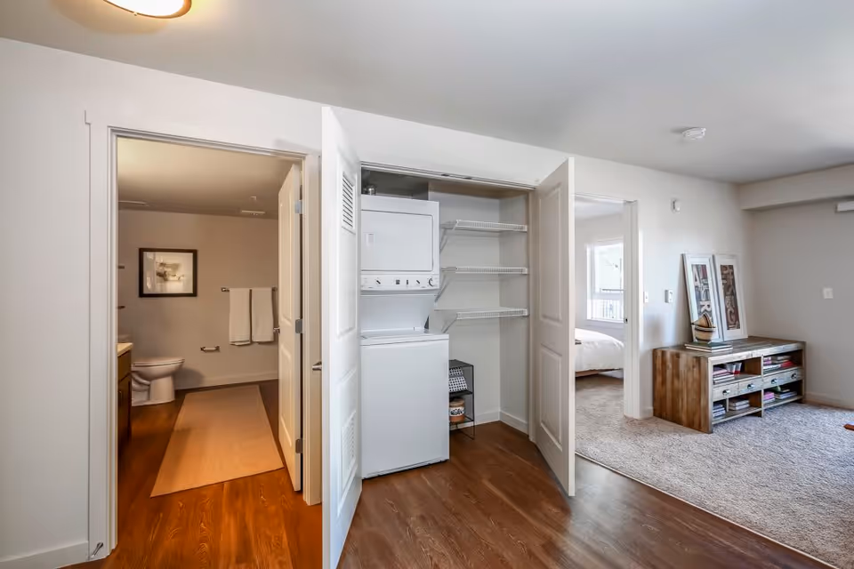 Interior view of an apartment showing a laundry closet with a stacked washer/dryer between a bathroom and a bedroom, with a living area and console table visible.