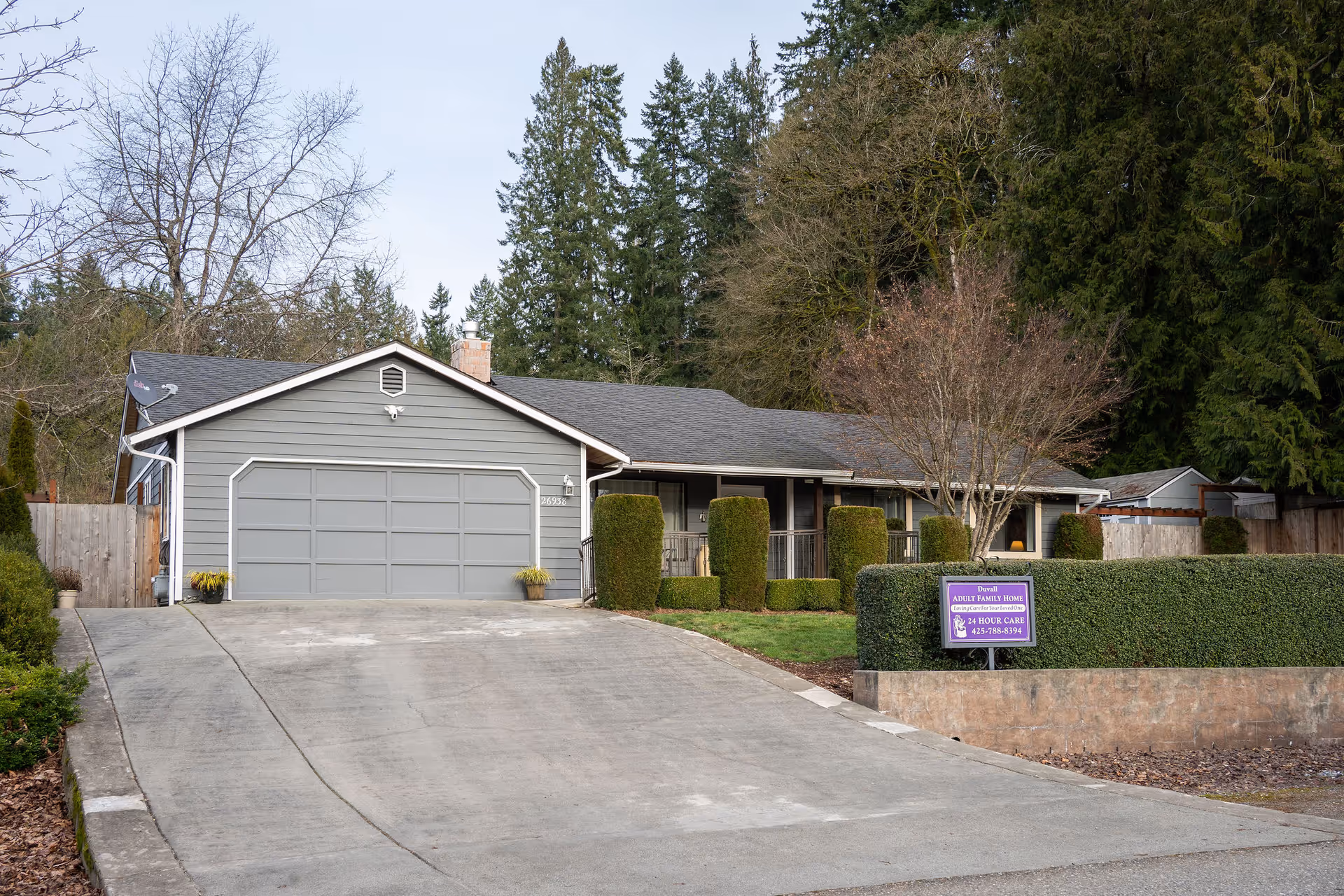 Front exterior of a single-story gray house with a two-car garage, driveway, trimmed hedges, and a purple sign for an adult family home.