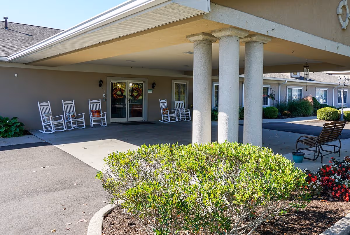Entrance area of Rolling Meadows Place featuring a covered drop-off with four white rocking chairs lined up against the building wall. There are double glass doors decorated with wreaths, surrounded by beige walls and windows. In front of the entrance, there is a landscaped area with green bushes and colorful flowers, and a bench with a small potted plant nearby.