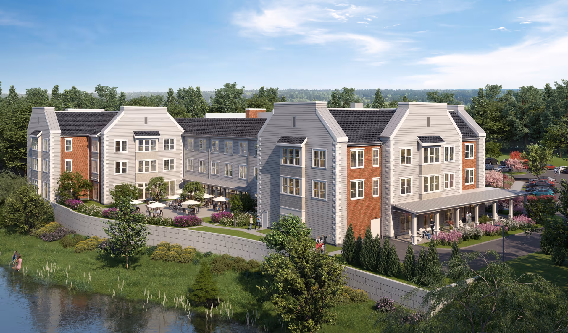 Exterior view of The Residence at Basking Ridge, a multi-story senior living facility with a mix of brick and light-colored siding. The building features multiple windows, a covered porch with seating, and an outdoor patio area with tables and umbrellas. The facility is surrounded by landscaped greenery, trees, and a small body of water in the foreground under a clear blue sky.