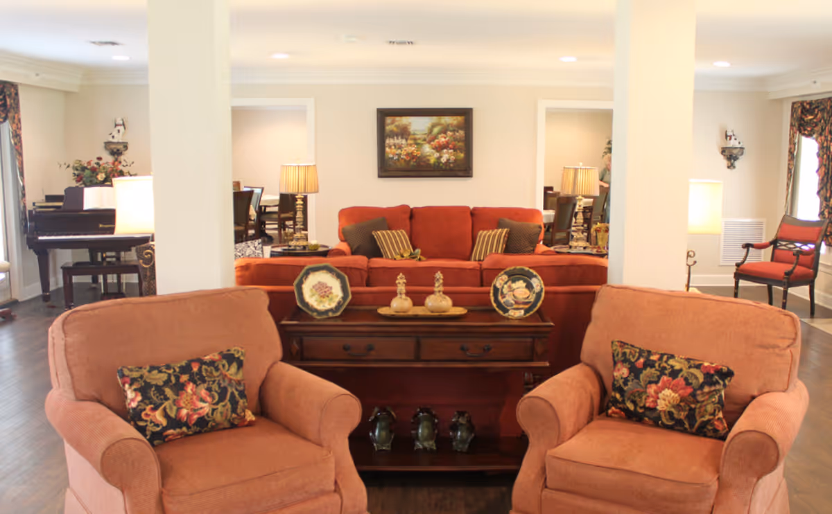Communal living room with two upholstered armchairs in front, a matching sofa, decorative console table with plates and ornaments, and a piano in the background.
