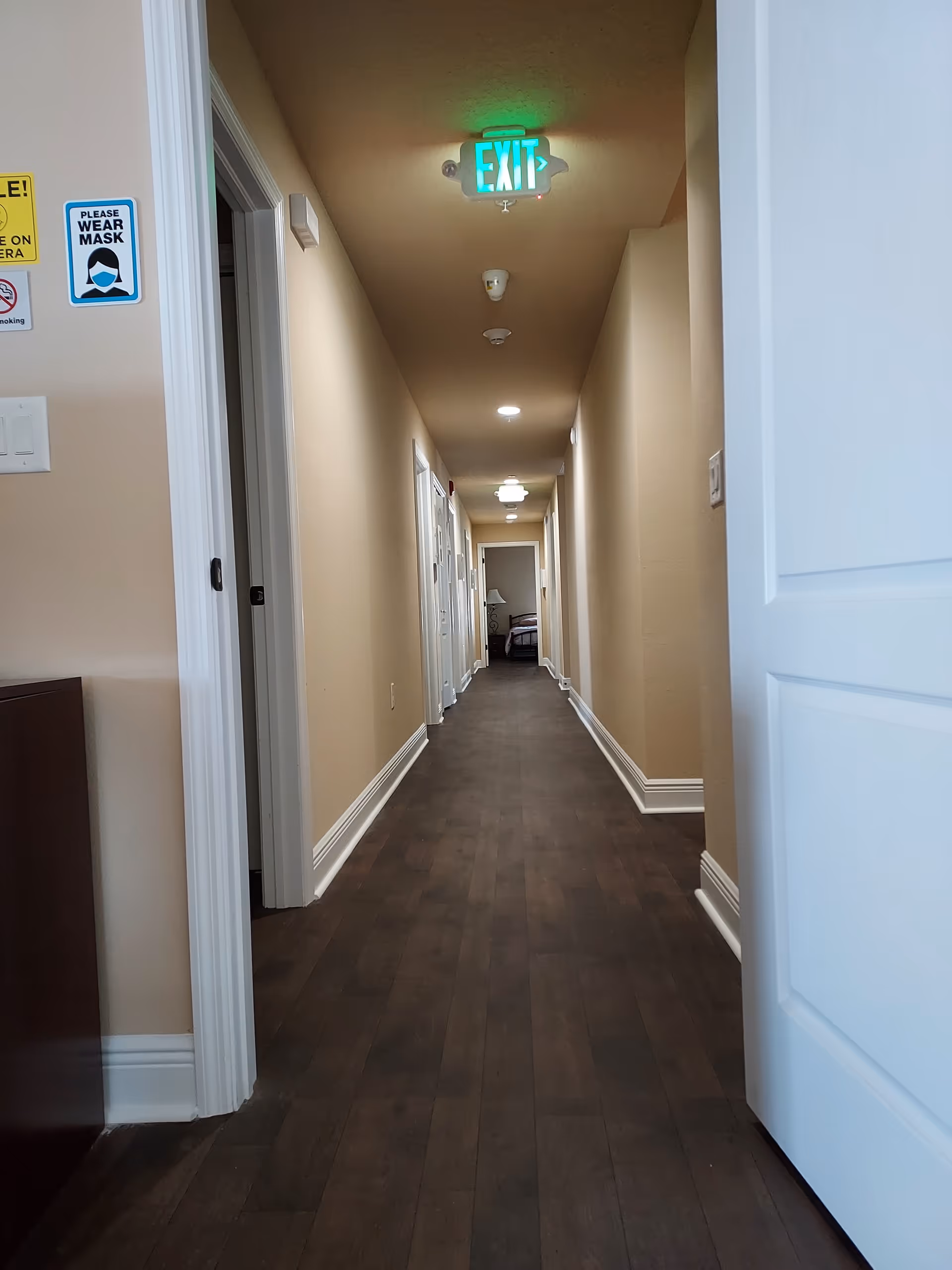 Long beige hallway in a senior living facility with doors and a green exit sign.