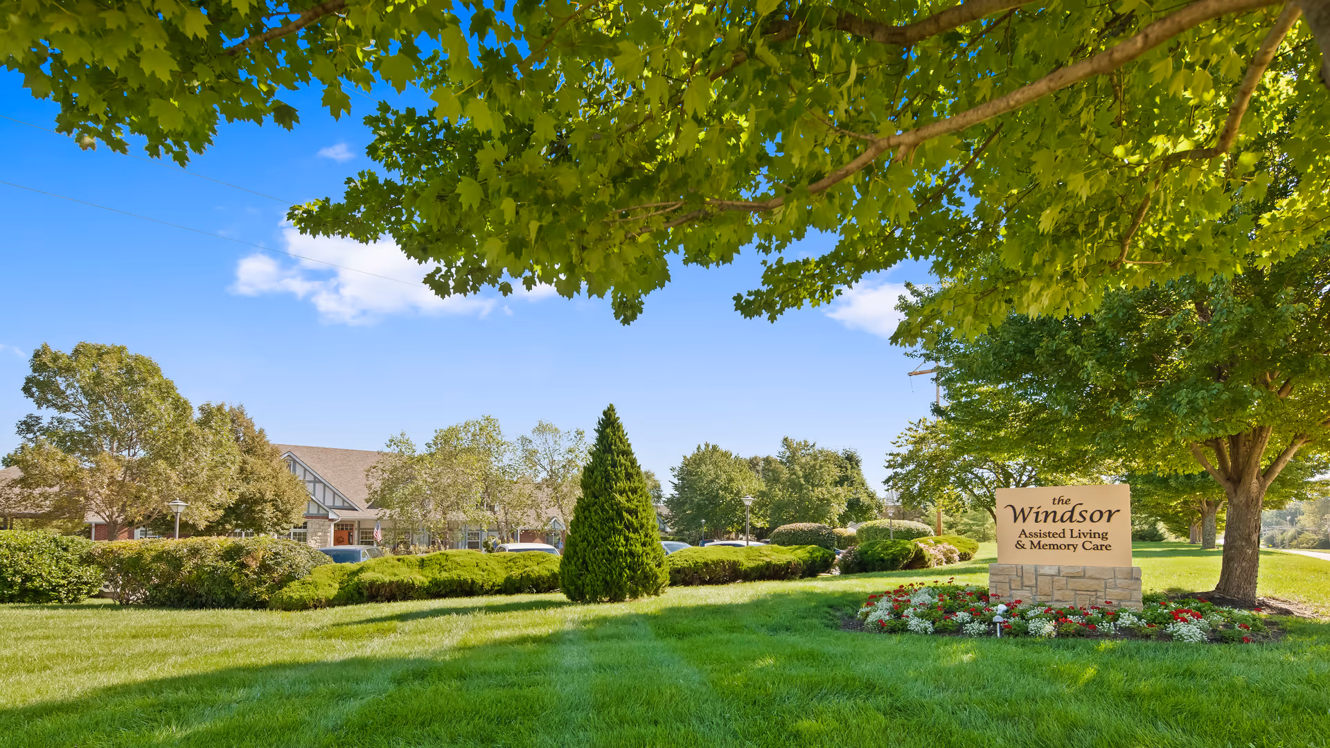 A sunny outdoor view of The Windsor of Lawrence assisted living and memory care facility, featuring a well-maintained lawn, trees, bushes, and a stone sign with flowers around it under a clear blue sky.