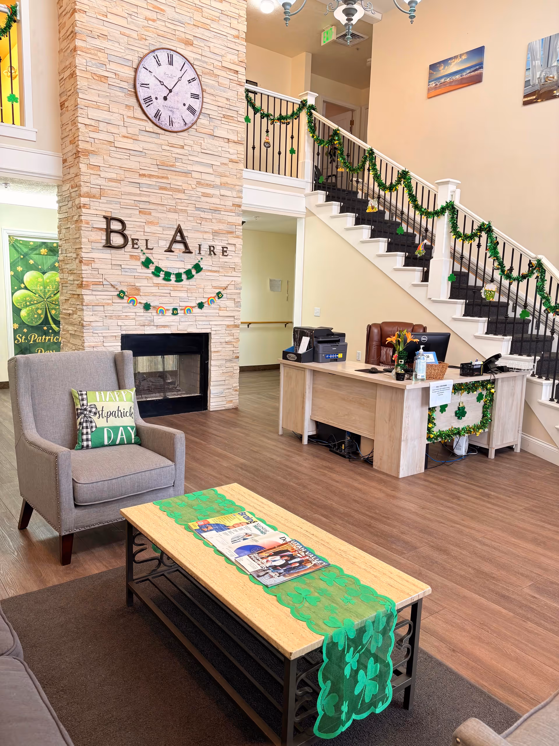 Interior view of a senior living facility lobby decorated for St. Patrick's Day with green shamrock garlands and banners. The room features a stone fireplace with a large clock above it, a comfortable armchair with a St. Patrick's Day themed pillow, a wooden coffee table with magazines and a green shamrock table runner, and a reception desk with a computer and office supplies. A staircase with black railings is decorated with green garlands.