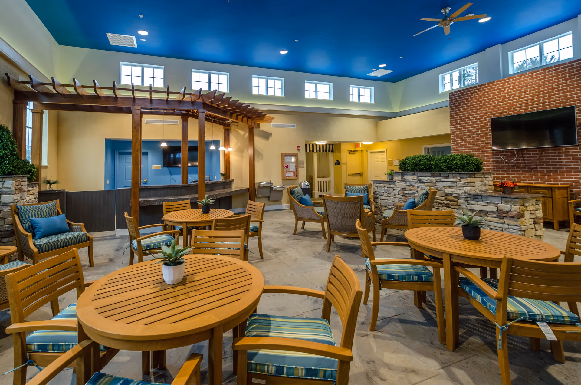A spacious indoor common area with multiple round wooden tables and chairs with blue striped cushions. There are also cushioned armchairs arranged near a stone partition with greenery on top. The room has a high ceiling painted blue with ceiling fans and several windows near the ceiling allowing natural light. A large flat-screen TV is mounted on a brick wall, and there is a wooden pergola structure near the back with pendant lights hanging above a counter area.