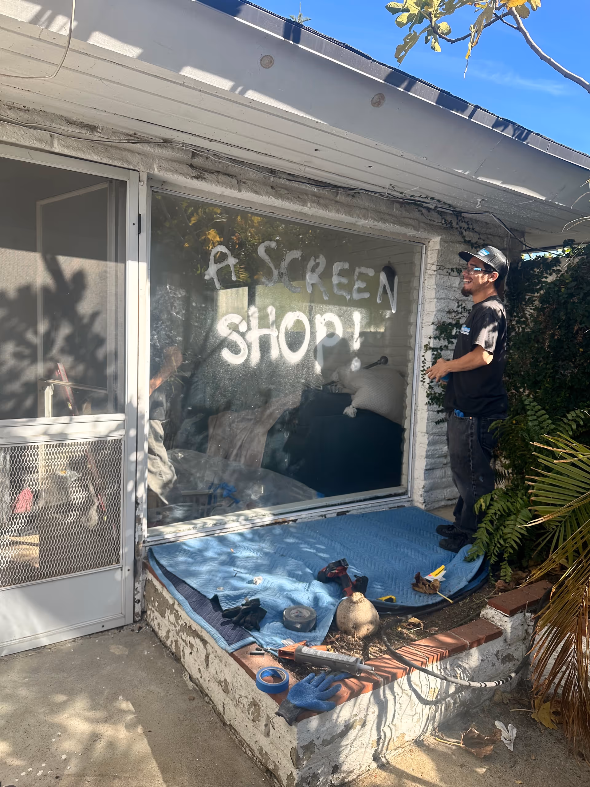 A man stands outside a storefront with graffiti on the window and tools on a tarp.