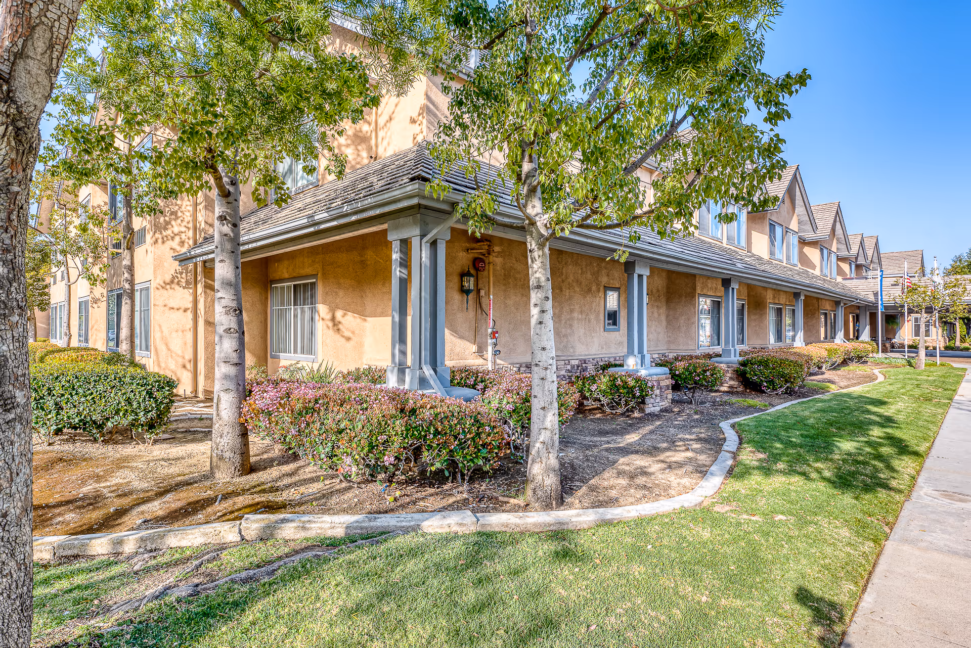 Front exterior of a two-story senior living building with a covered porch, landscaping, trees, and a sidewalk.