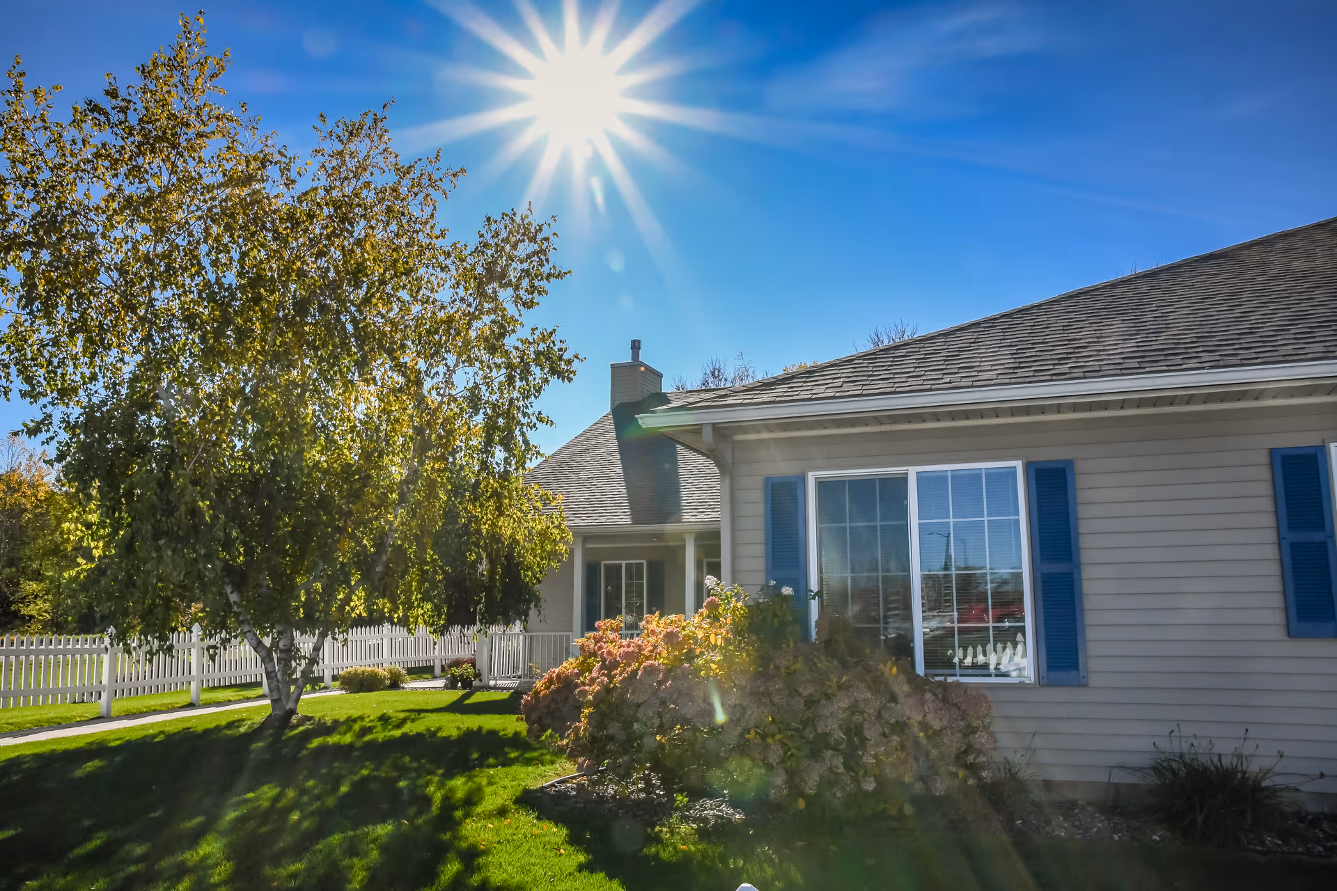 Front exterior of a single-story cottage with blue shutters, a white picket fence, lawn, tree and bright sun overhead.