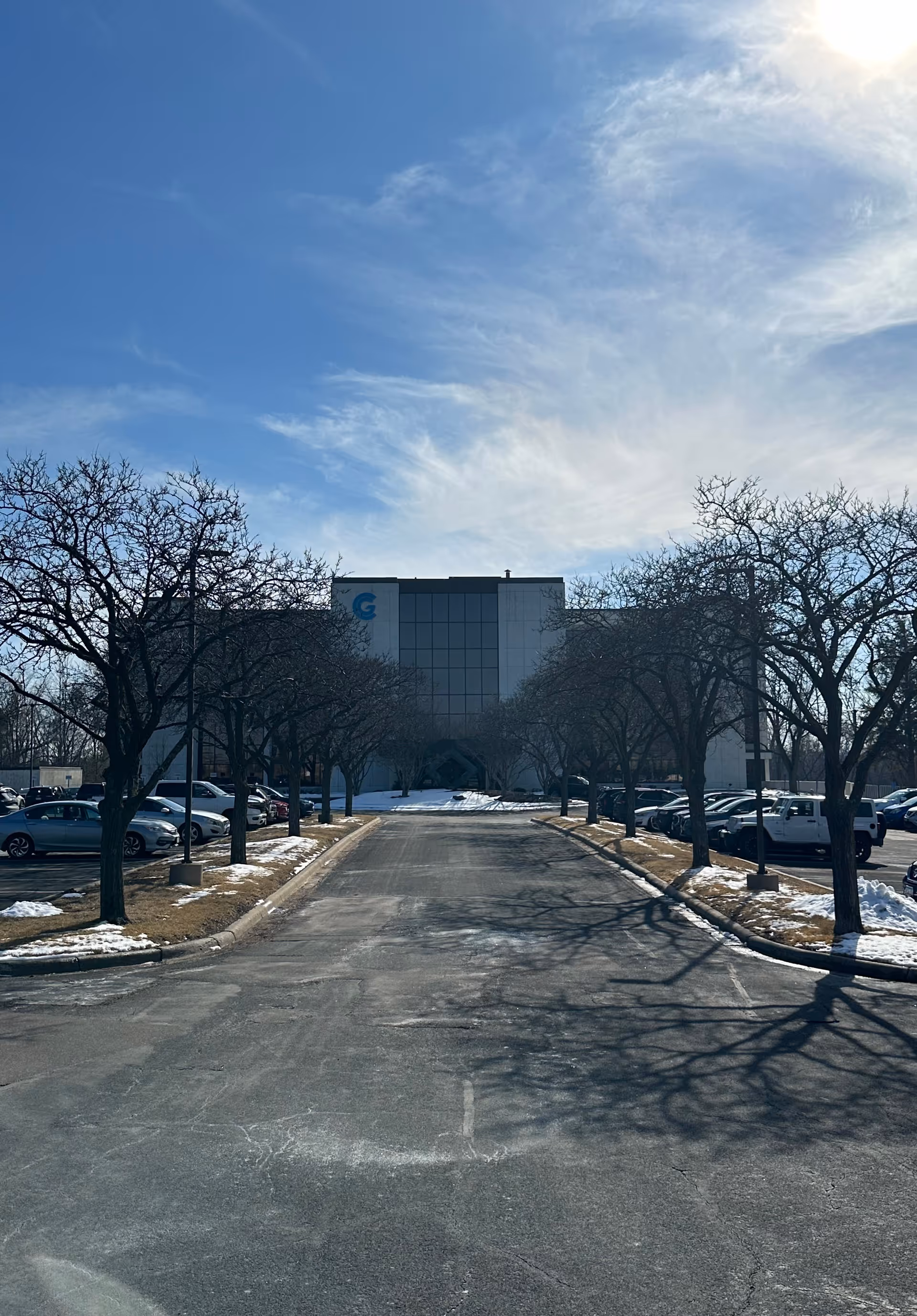 Winding driveway to a large building exterior, with bare trees and parked cars.