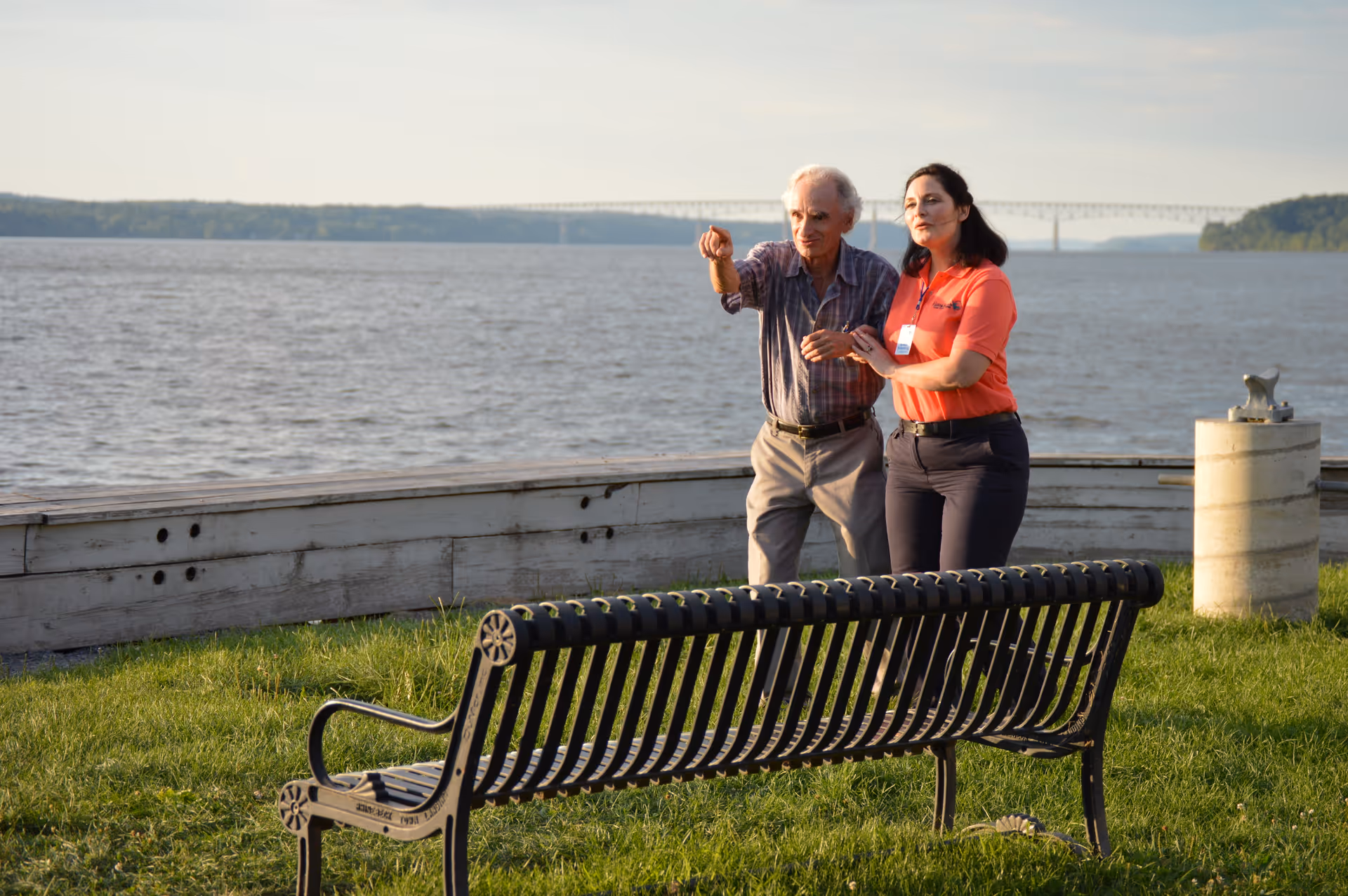 Elderly man and caregiver stand near a lakefront bench beside a bridge.