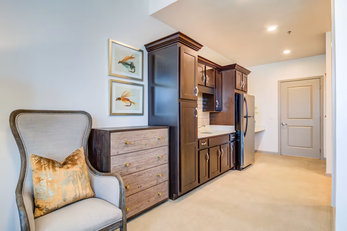 Interior view of a senior living facility kitchen area with dark wood cabinets, a stainless steel refrigerator, a light-colored countertop, and a beige carpeted floor. To the left, there is a light gray upholstered armchair with a gold decorative pillow and a wooden chest of drawers beneath two framed artworks of fishing flies on the wall.