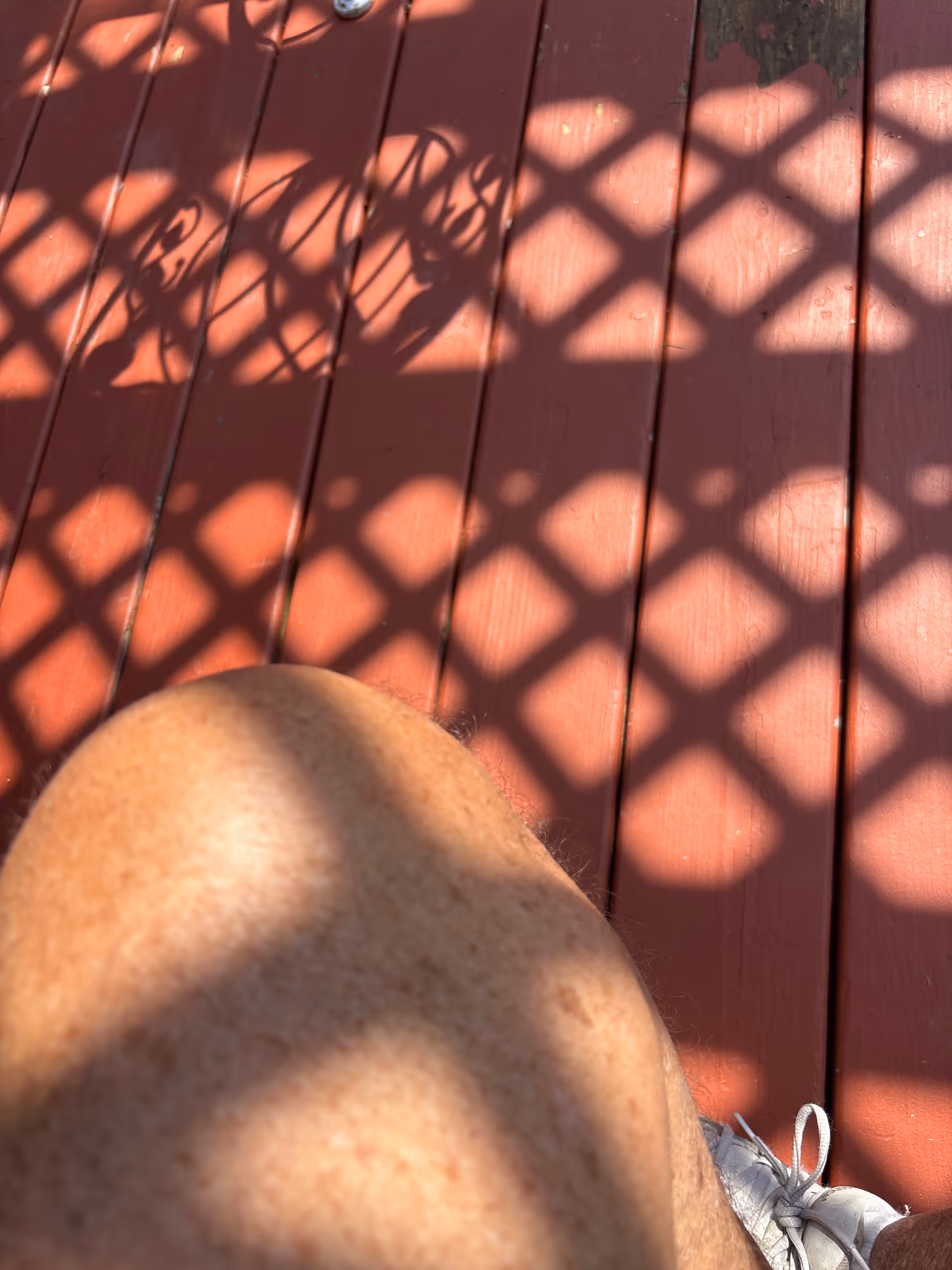 Red wooden deck with diamond lattice shadows and a person's knee in foreground.