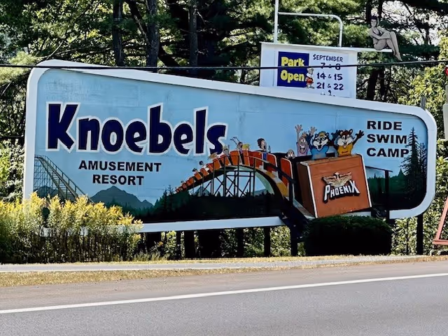 Outdoor road sign advertising Knoebels Amusement Resort with roller coaster illustration.