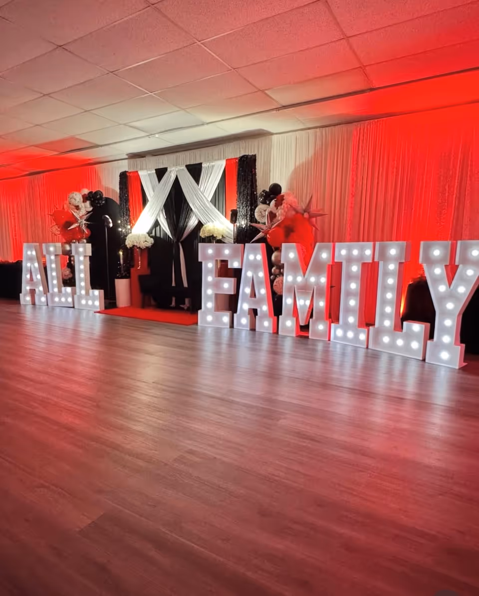 Indoor event space with a lit 'ALL FAMILY' sign, balloons, and elegant drapery backdrop.