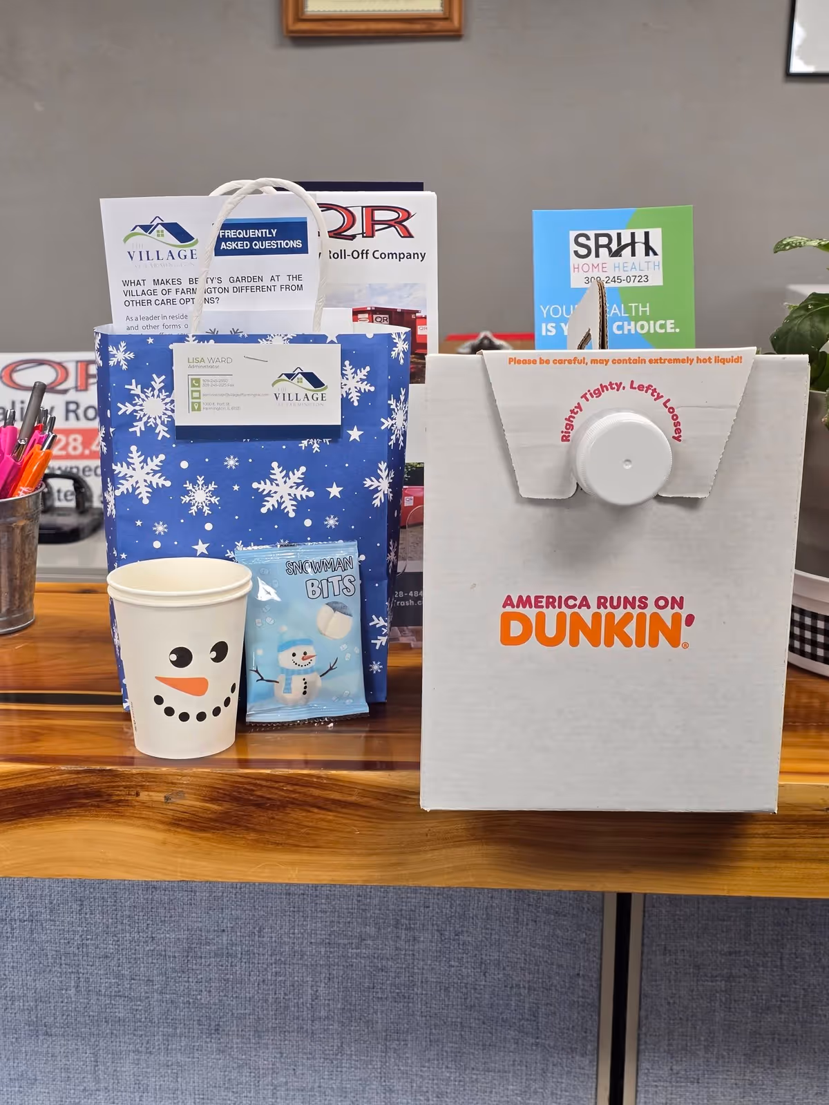 Indoor table display with holiday gift bags, snowman cup, and Dunkin box on wood surface.
