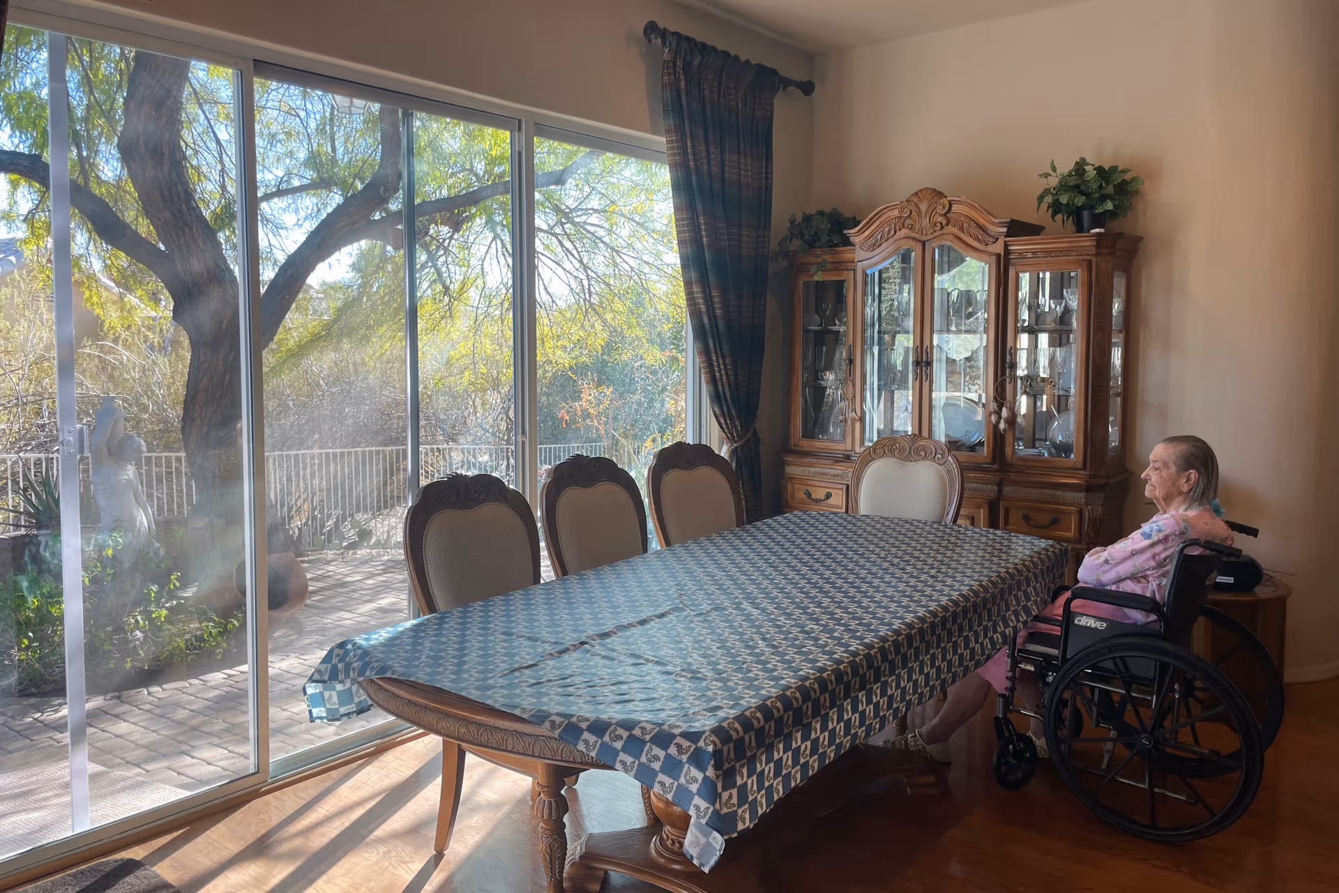 Sunlit dining room with a long table, china cabinet, and a resident in a wheelchair.