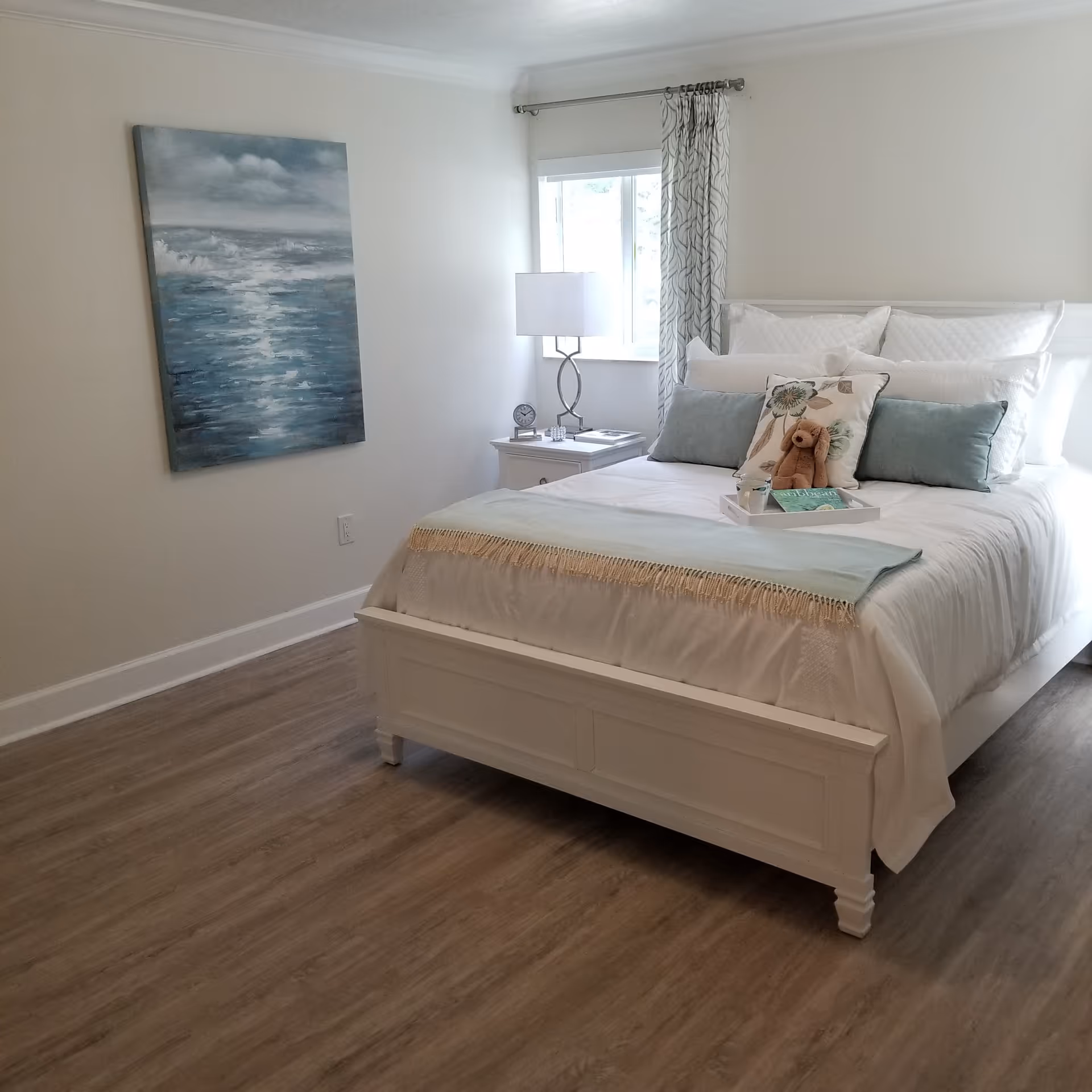 Bright bedroom with a white bed, nightstand, window, curtains, and coastal artwork.