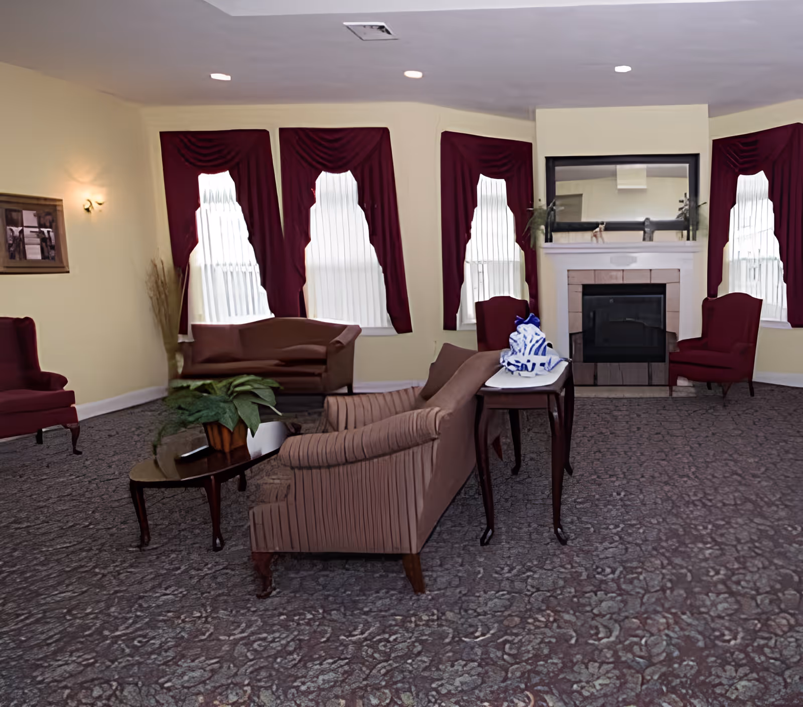 Lounge-style sitting room with sofas and armchairs arranged around a fireplace beneath windows with burgundy curtains.